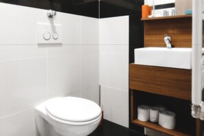 A modern bathroom with a white toilet, black and white tiled walls, a wooden shelf with toiletries, and a white sink. - Hotel Castle Rastatt (Rastatt)