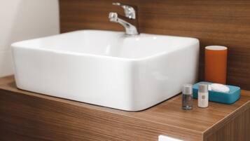 Ein weißes Waschbecken mit einem modernen Wasserhahn, einer Holzablage und einer kleinen blauen Seifenschale mit zwei Flaschen.