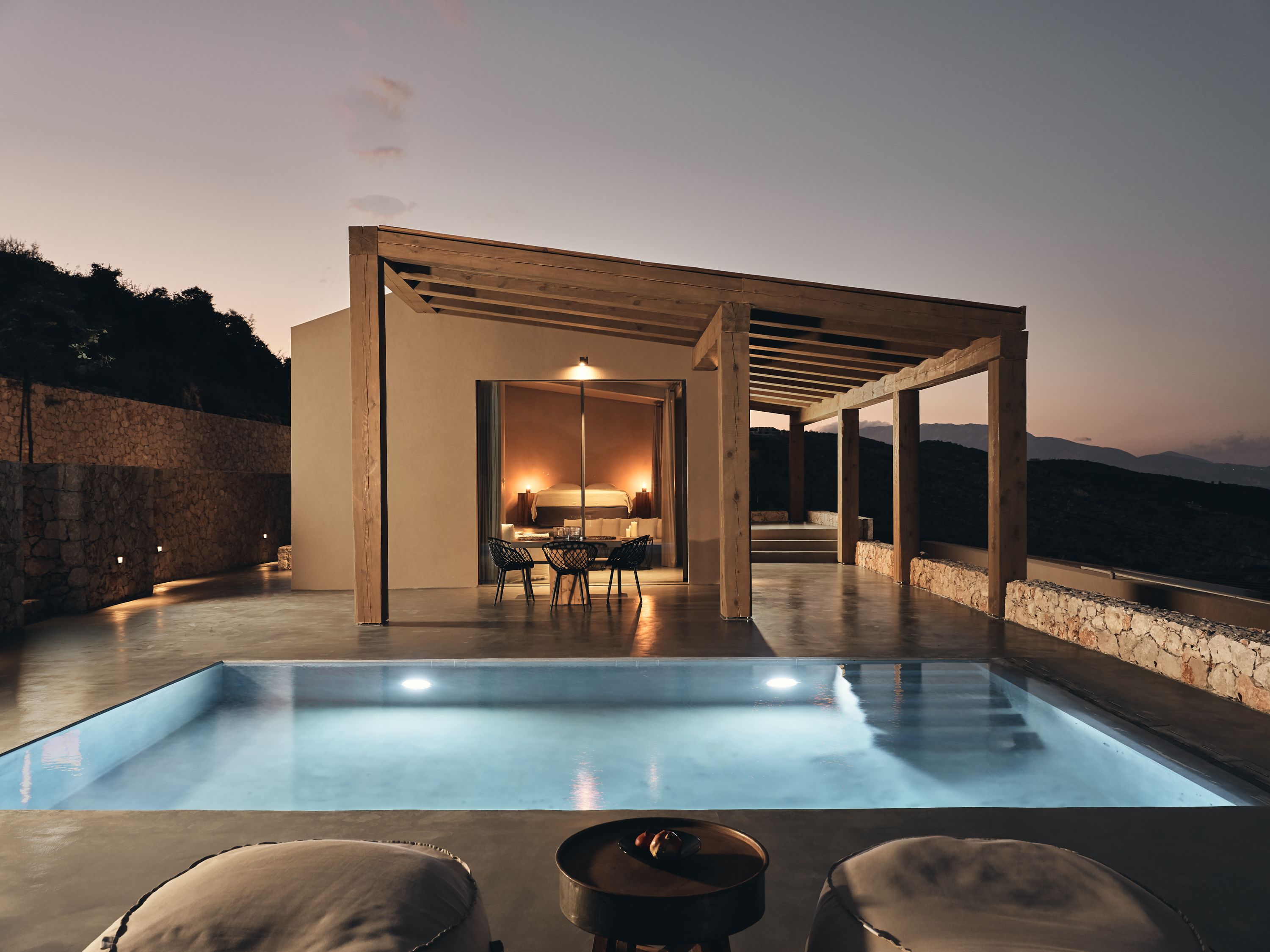 Un área de piscina moderna al aire libre con una pared de piedra, una pérgola de madera y vistas a las montañas al fondo.