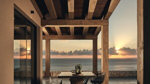 A modern outdoor dining area with a view of the ocean at sunset, featuring a wooden pergola, a table with chairs, and a bench. - Vais luxury villas (Zakynthos)