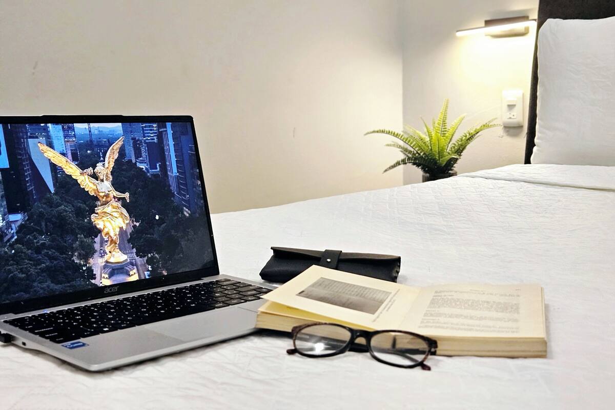 A laptop on a bed with a book and glasses beside it, a small plant, and a night scene on the laptop screen.