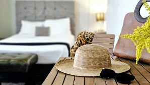 A room with a bed, a wooden table with a straw hat and black sunglasses, and a leopard print throw. - Singular Goldsmith (Mexico City)