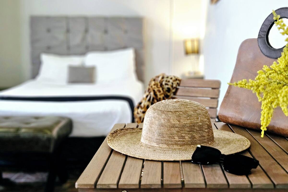A room with a bed, a wooden table with a straw hat and black sunglasses, and a leopard print throw.