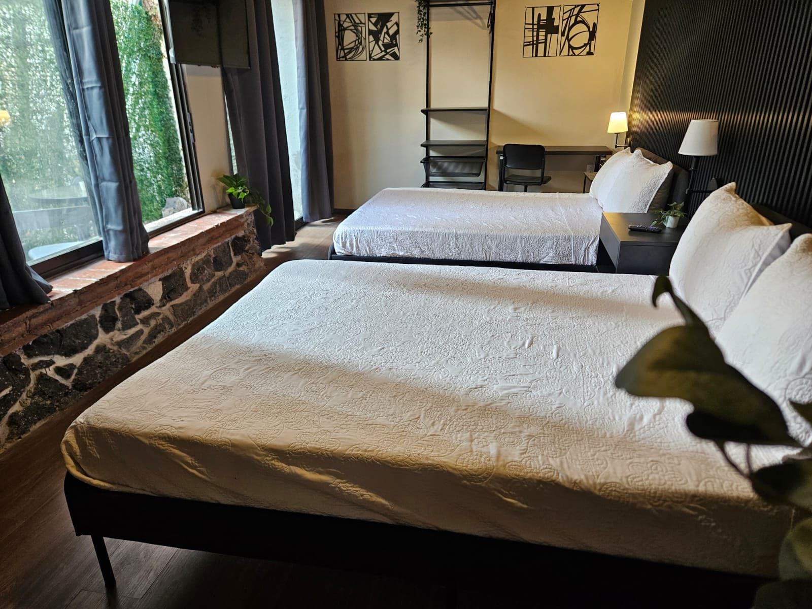 A hotel room with two beds, a stone wall, a desk with a lamp, and artwork on the wall.