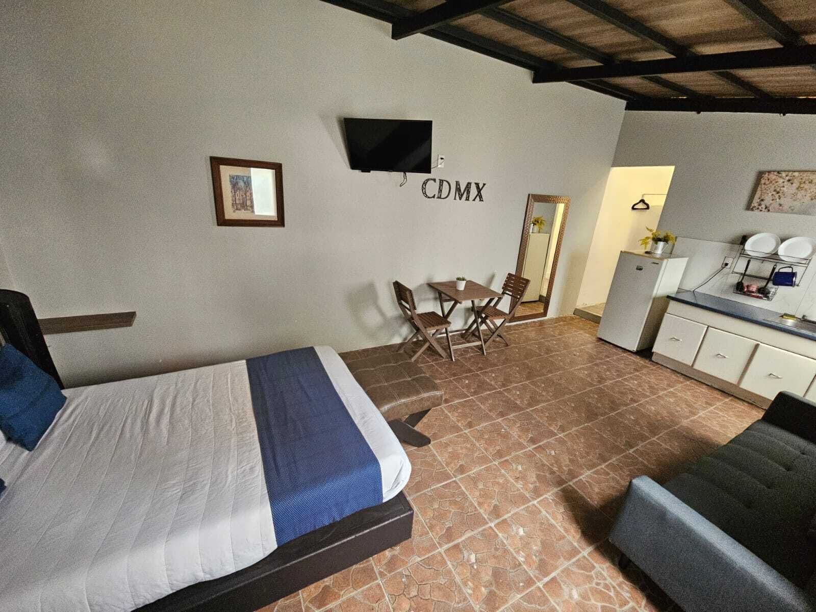 A bedroom with a bed, a small dining table with chairs, a kitchenette, and a television mounted on the wall.