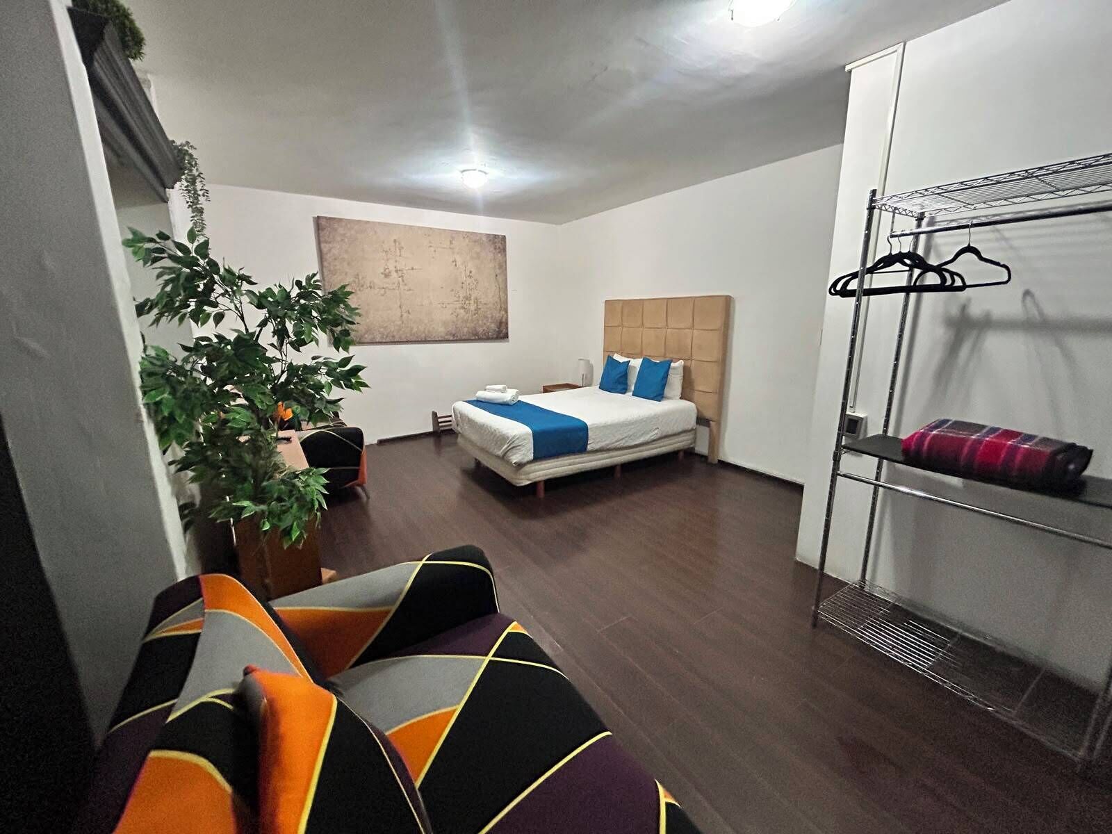 A hotel room with a bed, a seating area with a sofa, a plant, a wall-mounted artwork, and a wardrobe with hanging items.