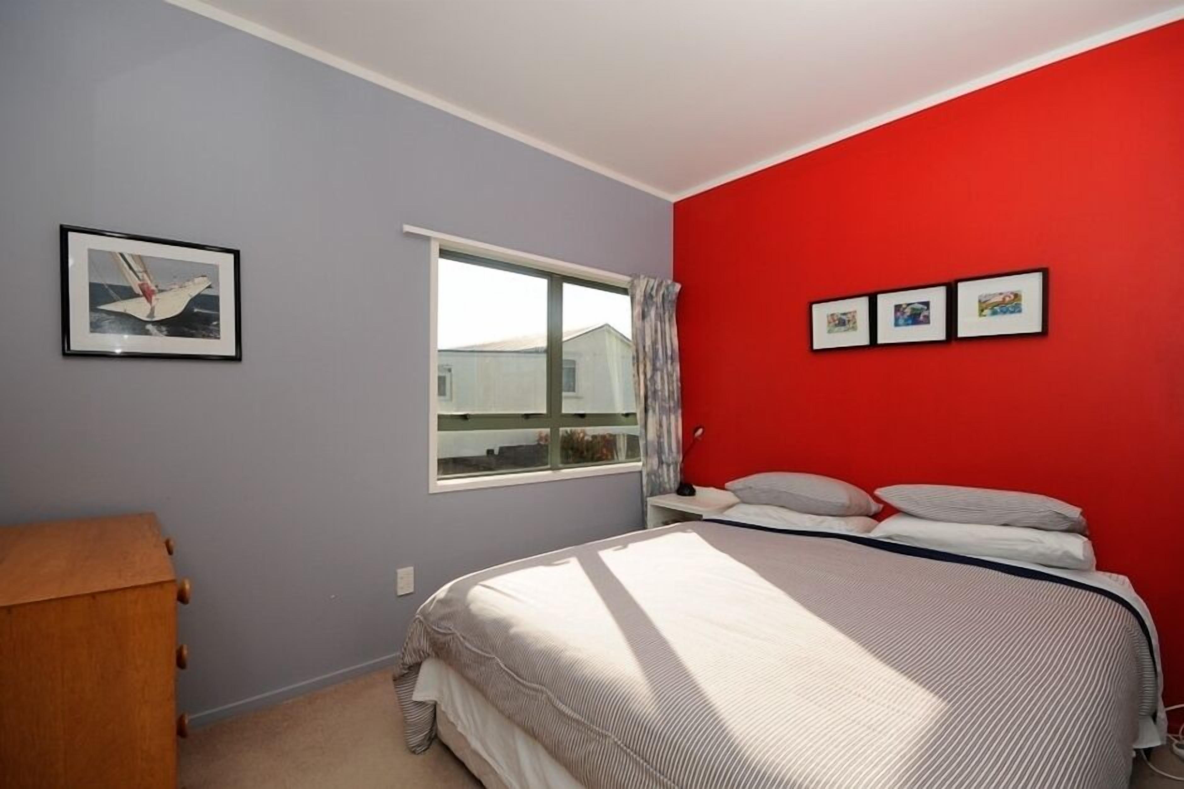 A bedroom with a bed, a window, a wooden dresser, and a red wall.