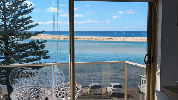 Un balcón con puerta de cristal, sillas blancas y vista a la playa y al océano.