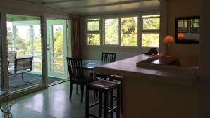 Quarto com área de refeições, bancada de cozinha e vista para árvores através de grandes janelas.