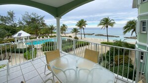 Een balkon met een glazen tafel en stoelen, met uitzicht op een zwembad, palmbomen en een strand.