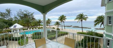 Un balcon avec une table et des chaises en verre, donnant sur une piscine, des palmiers et une plage.