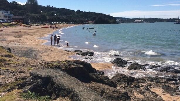 Une plage au rivage rocheux, où l’on voit des gens se promener et nager, et où l’on aperçoit une ville au loin.