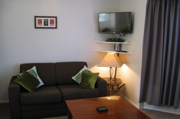 A living room with a sofa, a coffee table, a TV on a stand, a lamp, and a picture frame on the wall. - Koru Cottage - Pristine and newly renovated. (Paihia)