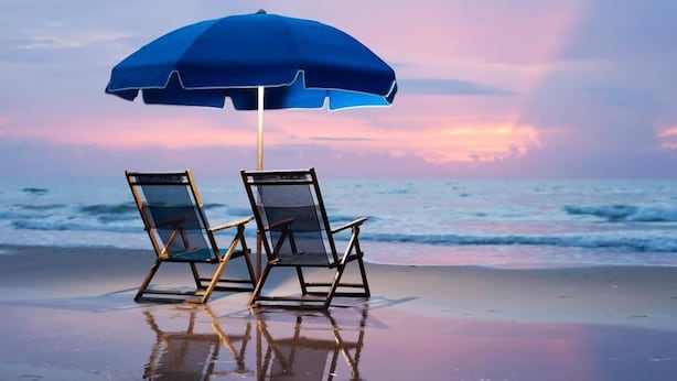 Zwei Liegestühle unter einem blauen Sonnenschirm an einem sandigen Strand, im Hintergrund ein Sonnenuntergang.