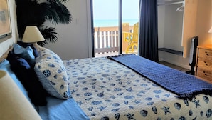 Ein Schlafzimmer mit Bett, Nachttisch, Lampe, Kommode und Meerblick durch eine Schiebetür.