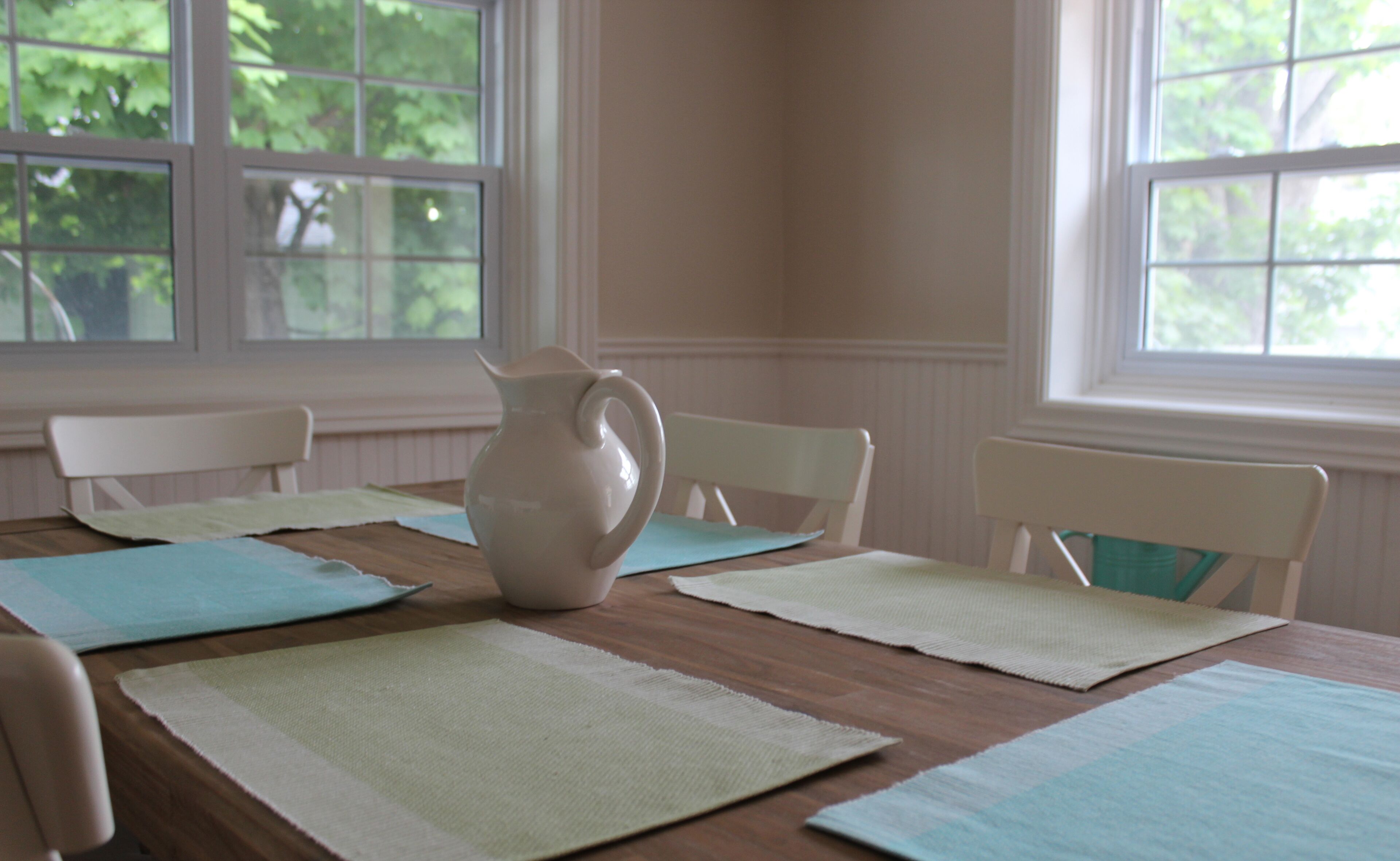 Une table à manger avec des sets de table, un pichet et des chaises blanches, dans une pièce dotée de fenêtres et offrant une vue sur les arbres.