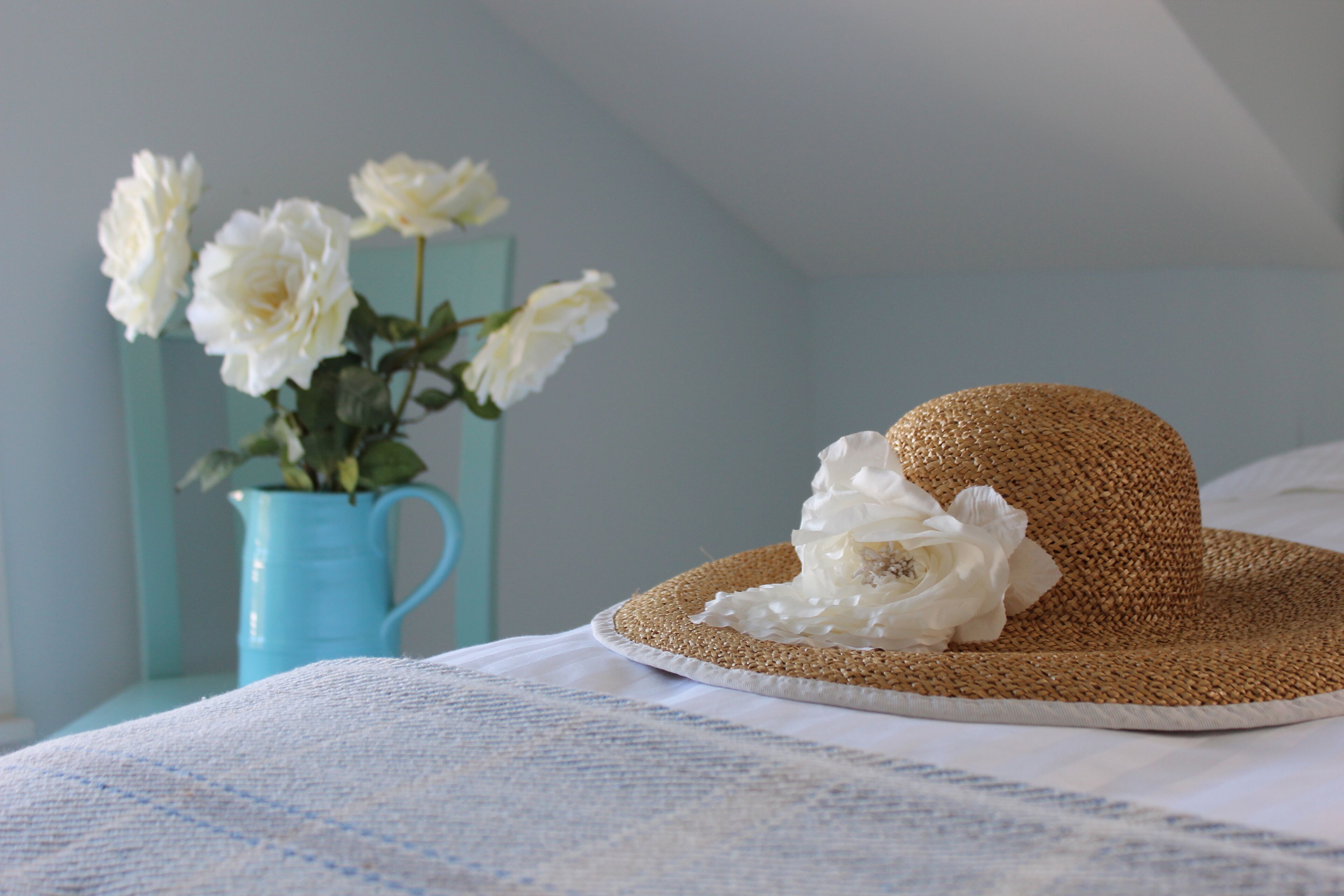 Ein Strohhut mit weißen Blumen auf einem Bett mit einer blau-weißen karierten Tagesdecke.