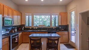A modern kitchen with wooden cabinets, a central island with two chairs, a stove, and a large window providing a view of trees. - Whole Home Air Cleaner, Quiet surroundings, deck and Jacuzzi, near golf course (Austin)