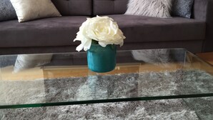 A glass coffee table with a teal vase containing white flowers, positioned in front of a dark grey sofa with a fluffy pillow and a grey knitted throw. - Whole Home Air Cleaner, Quiet surroundings, deck and Jacuzzi, near golf course (Austin)
