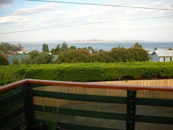 A view from a balcony with a wooden railing, overlooking a body of water, greenery, and distant buildings.