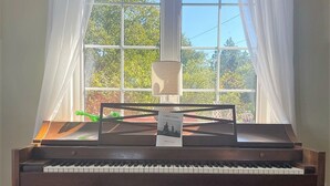 A grand piano in a room with a large window, a lamp, and a framed picture on the piano. - Breathtaking Remote Oceanfront Oasis Panoramic Clifftop Views (Lubec)