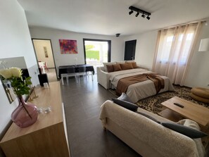 A modern living room with a sofa, a dining area with a table and chairs, a bed with a brown blanket, and a large window with curtains.
