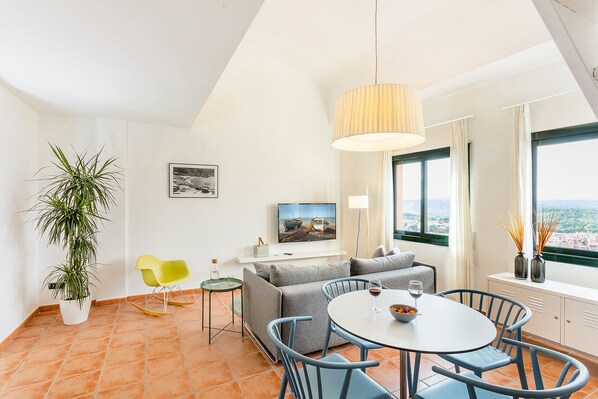 A modern living room with a sofa, dining table, and chairs, a TV, a plant, and a view of the city through large windows. - Begur Sunset ,2 bedroom loft close to the center of Begur. With terrace and gorgeous sea views (H42) (Begur)