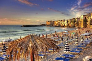 A beach with thatched umbrellas and sun loungers, a pier, and historic buildings along the shore.