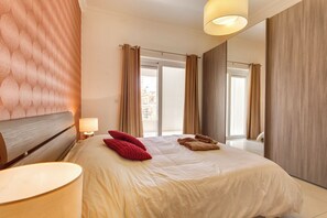 A hotel room with a large bed, a bedside lamp, a view of the city, and a window with curtains.