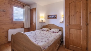 Ein Zimmer mit Holzboden, einem Bett, einem Fenster mit Vorhängen, einer Tür und einer Wandlampe.