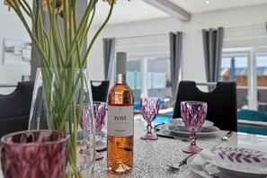 Uma mesa de jantar com uma garrafa de vinho, copos de vidro rosa e um vaso com hastes verdes.