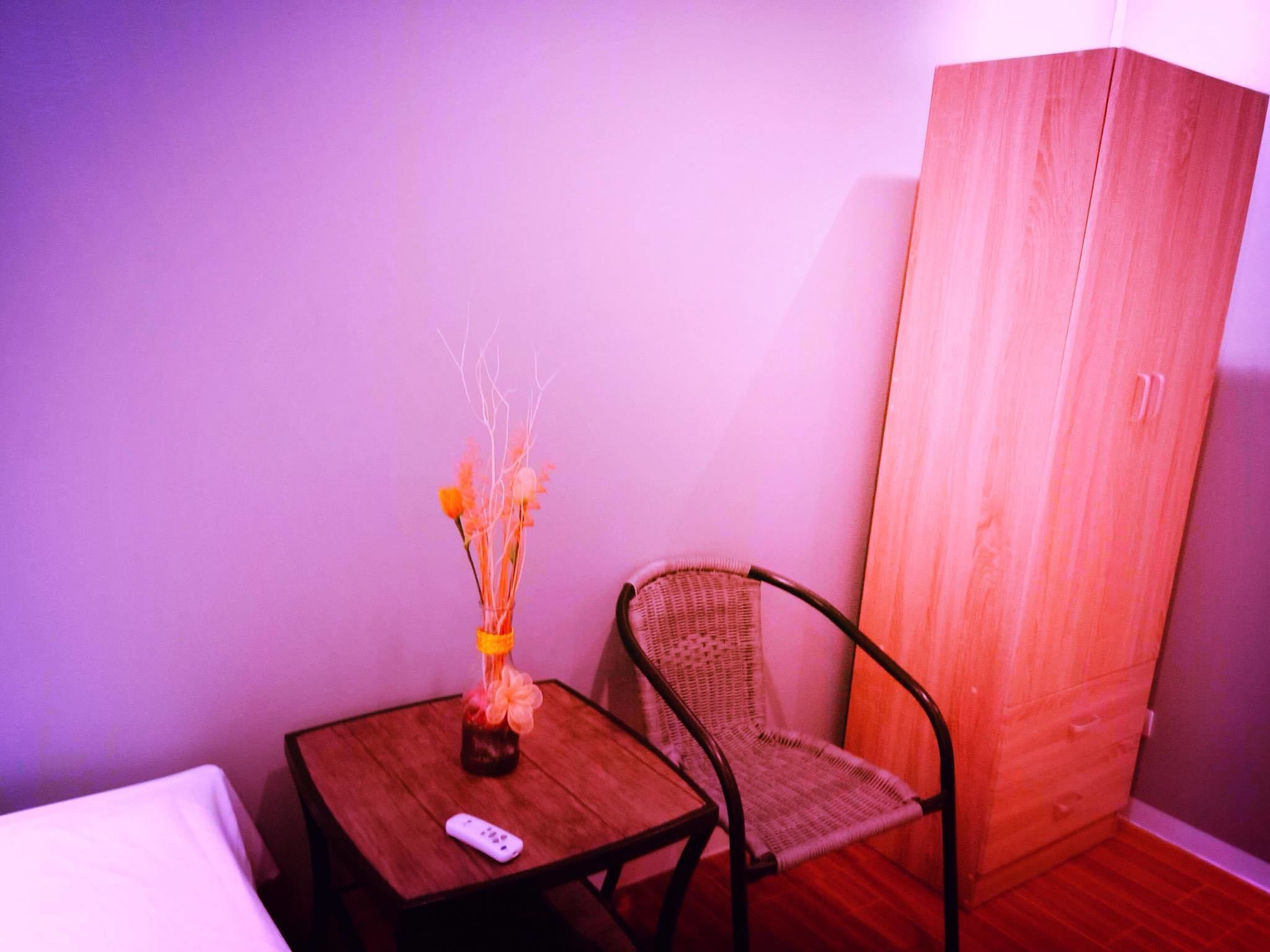 A small room with a wooden cabinet, a wicker chair, a small table with a vase and a remote control.