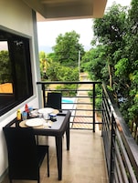A balcony with a table set for dining, a pool in the background, and lush greenery surrounding the area.