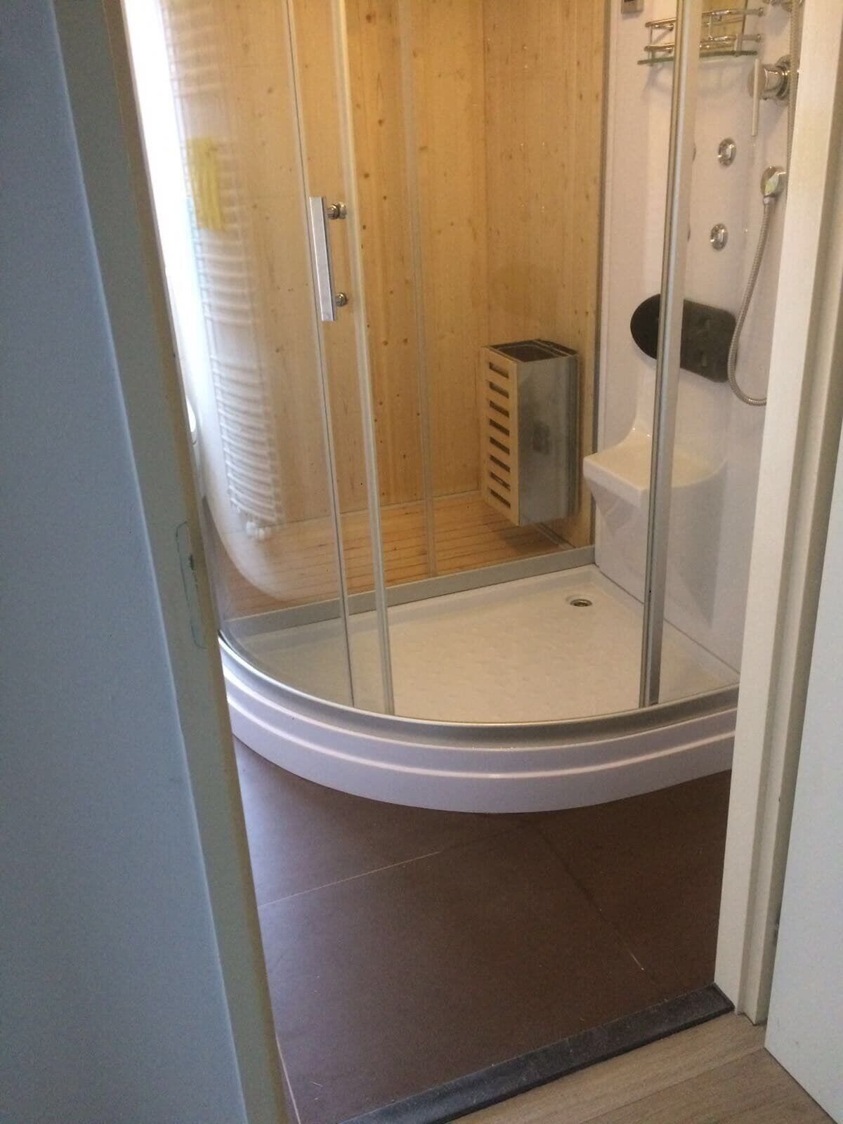 Une cabine de douche fermée par des parois en verre, avec un sol blanc, une porte en bois et une poubelle en métal.