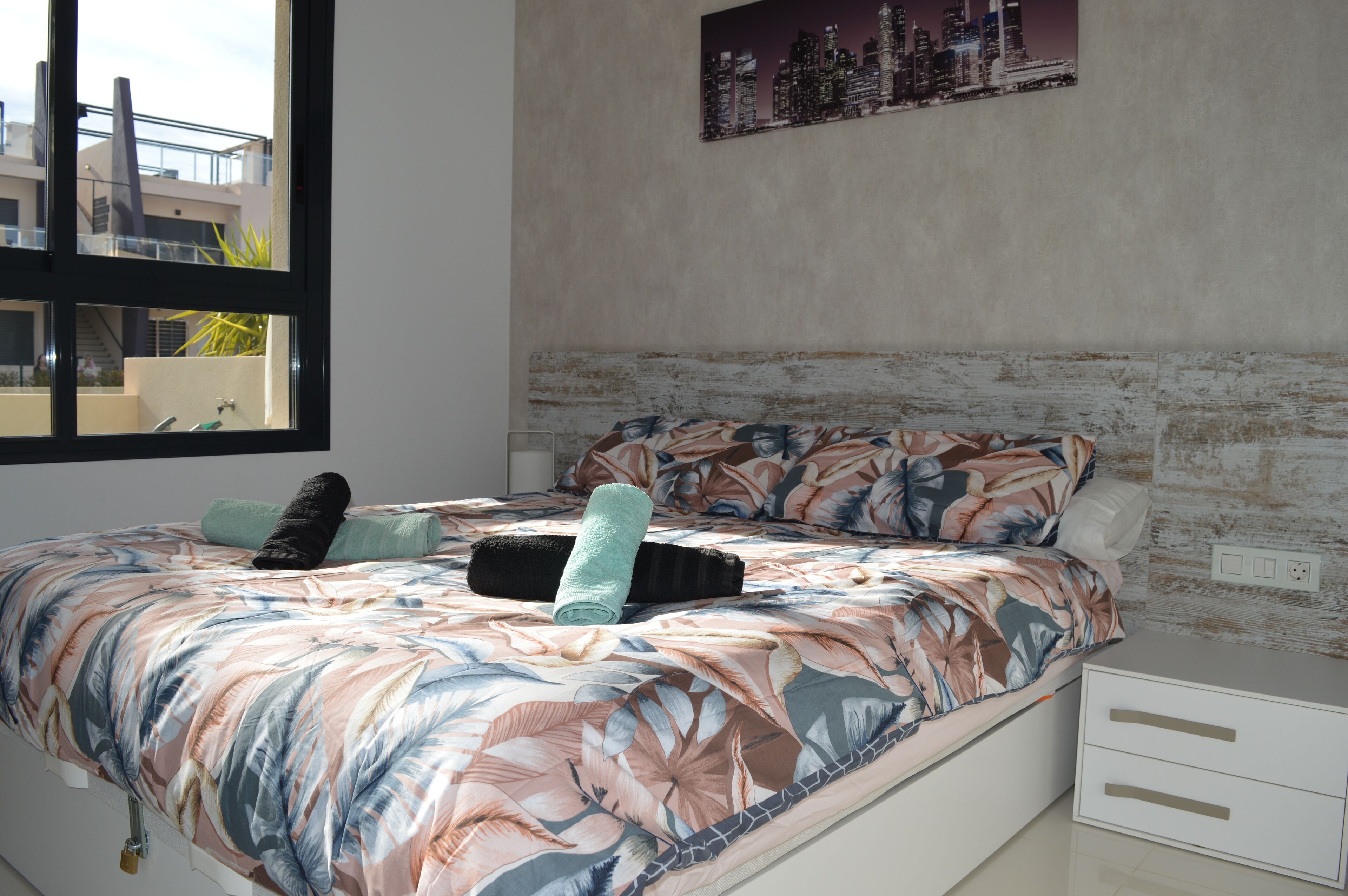 Une chambre à coucher avec un lit, une table de chevet, une fenêtre et un tableau représentant un paysage urbain accroché au mur.