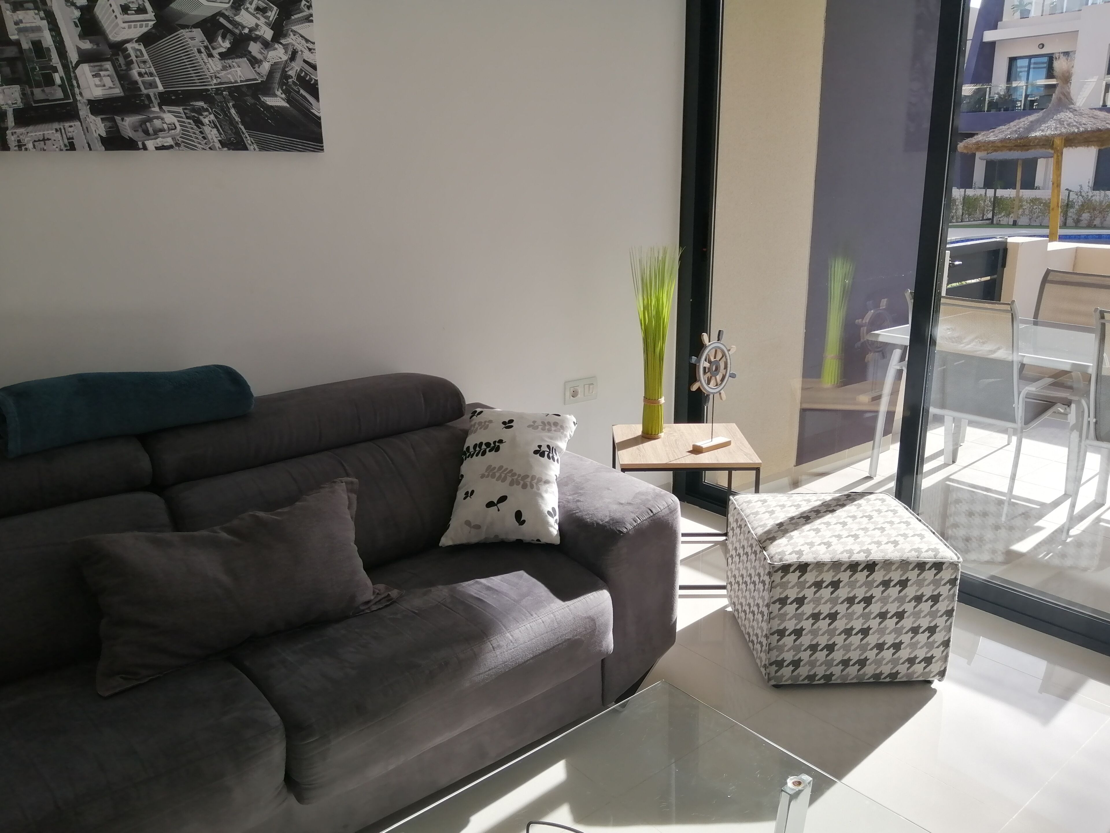 Un salon moderne avec un canapé gris, une table basse en verre et un pouf à motifs. On y trouve une petite table d’appoint sur laquelle est posé un vase contenant des plantes vertes.