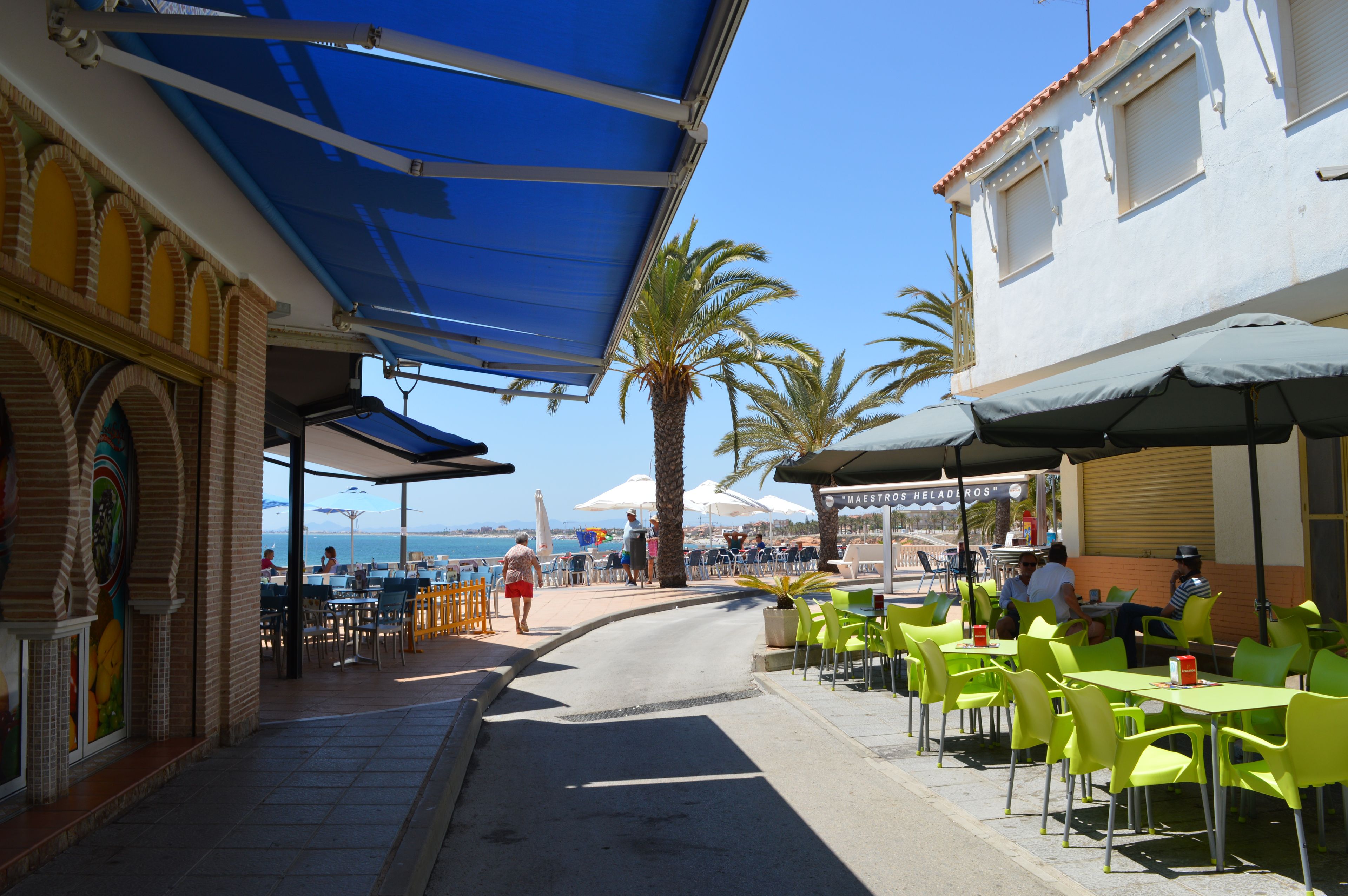 An outdoor cafe with tables and chairs, a blue awning, and a view of the sea.