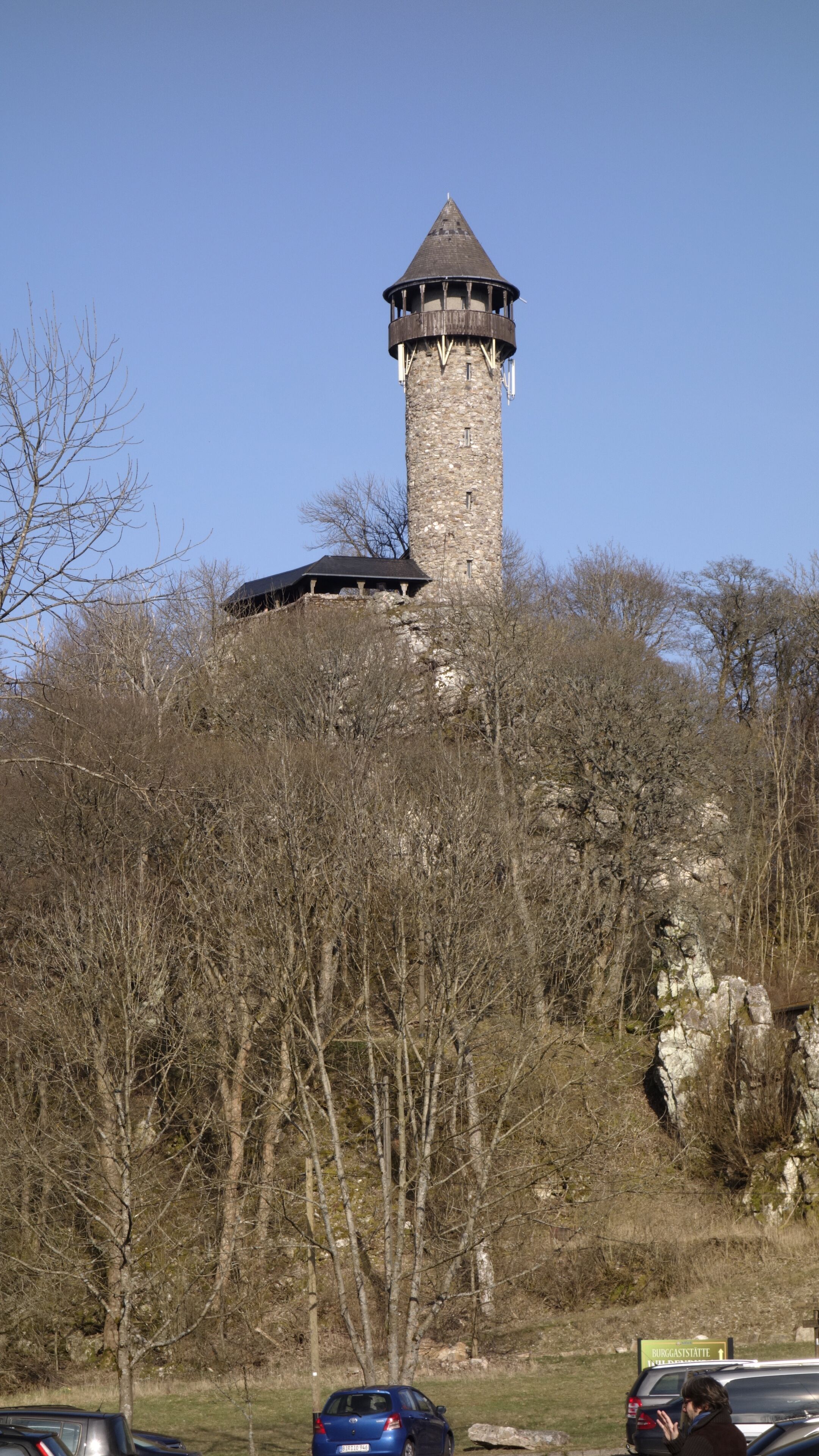 Een stenen toren met een puntdak, omgeven door bomen en een parkeerplaats.