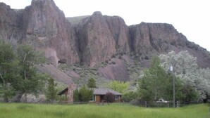 A rustic cabin with a stone chimney, surrounded by a grassy field and a forested mountain backdrop.