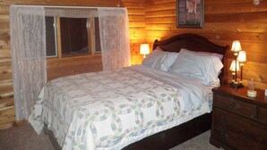 A wooden cabin bedroom with a bed, two bedside lamps, a window with curtains, and a framed picture on the wall.