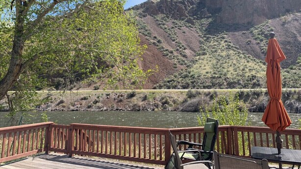 A wooden deck with chairs and an umbrella, overlooking a river and a mountainous landscape.