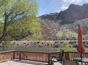 Une terrasse en bois, dotée de chaises et d’un parasol, qui offre une vue sur une rivière et un paysage montagneux.