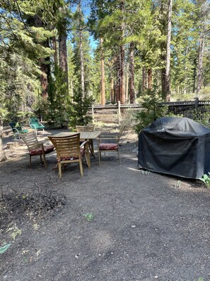 Un espace extérieur aménagé avec des tables et des chaises en bois, situé dans un cadre boisé, et doté d’un barbecue couvert.