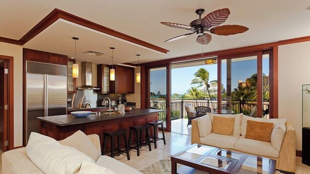 A modern kitchen with a central island, a dining area with a sofa and chairs, and a balcony with a view of palm trees. - Exceptional 5-stars Beach Villas at Ko Olina - 3 Bedrooms & 3 Full Bathrooms (Ko Olina)