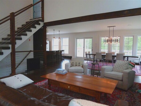 A spacious living area with a wooden coffee table, a sofa, and a dining table with chairs. A staircase is visible in the background.