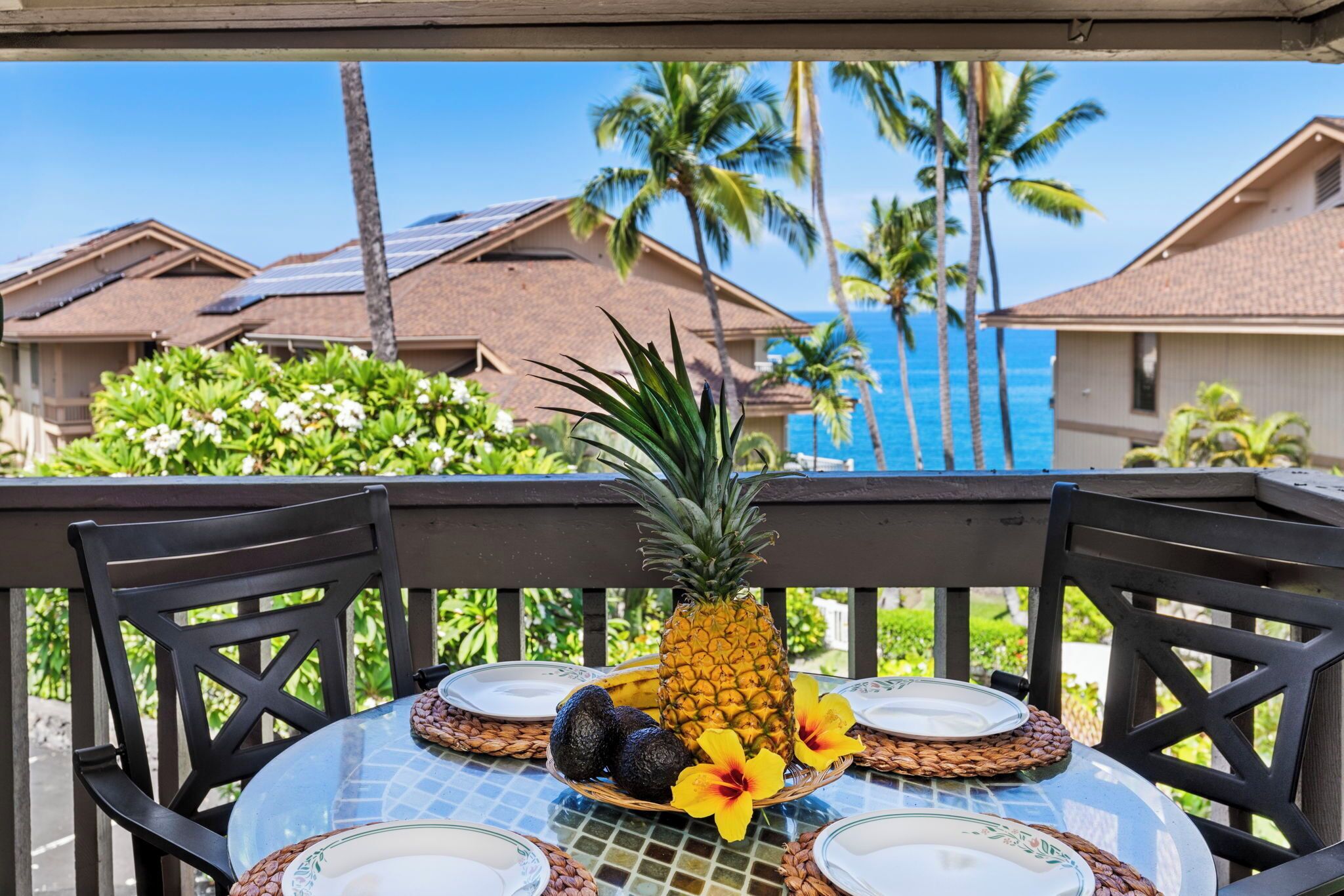 A table with a pineapple, two avocados, and a yellow flower on a woven placemat, set on a balcony with a view of palm trees and houses.