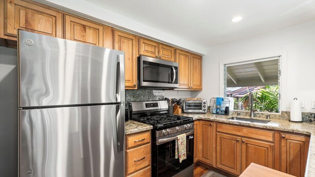 A kitchen with wooden cabinets, stainless steel appliances, a microwave, a stove, and a window with a view of greenery.