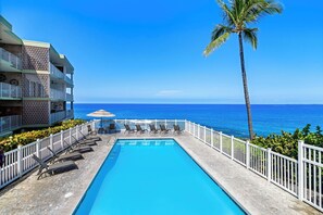 A swimming pool with lounge chairs and umbrellas, surrounded by a white fence, with a view of the ocean and a palm tree. - Spacious 2BR/2BA Condo - Ocean View, Above it All, Oceanside Pool and 3 Zone A/C (Kailua-Kona)