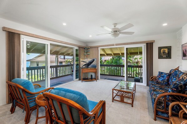 A spacious living room with a glass door leading to a balcony, a ceiling fan, and a television on a stand. - Spacious 2BR/2BA Condo - Ocean View, Above it All, Oceanside Pool and 3 Zone A/C (Kailua-Kona)