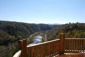 Une terrasse en bois donnant sur une rivière, avec une vallée boisée et des montagnes lointaines en toile de fond.